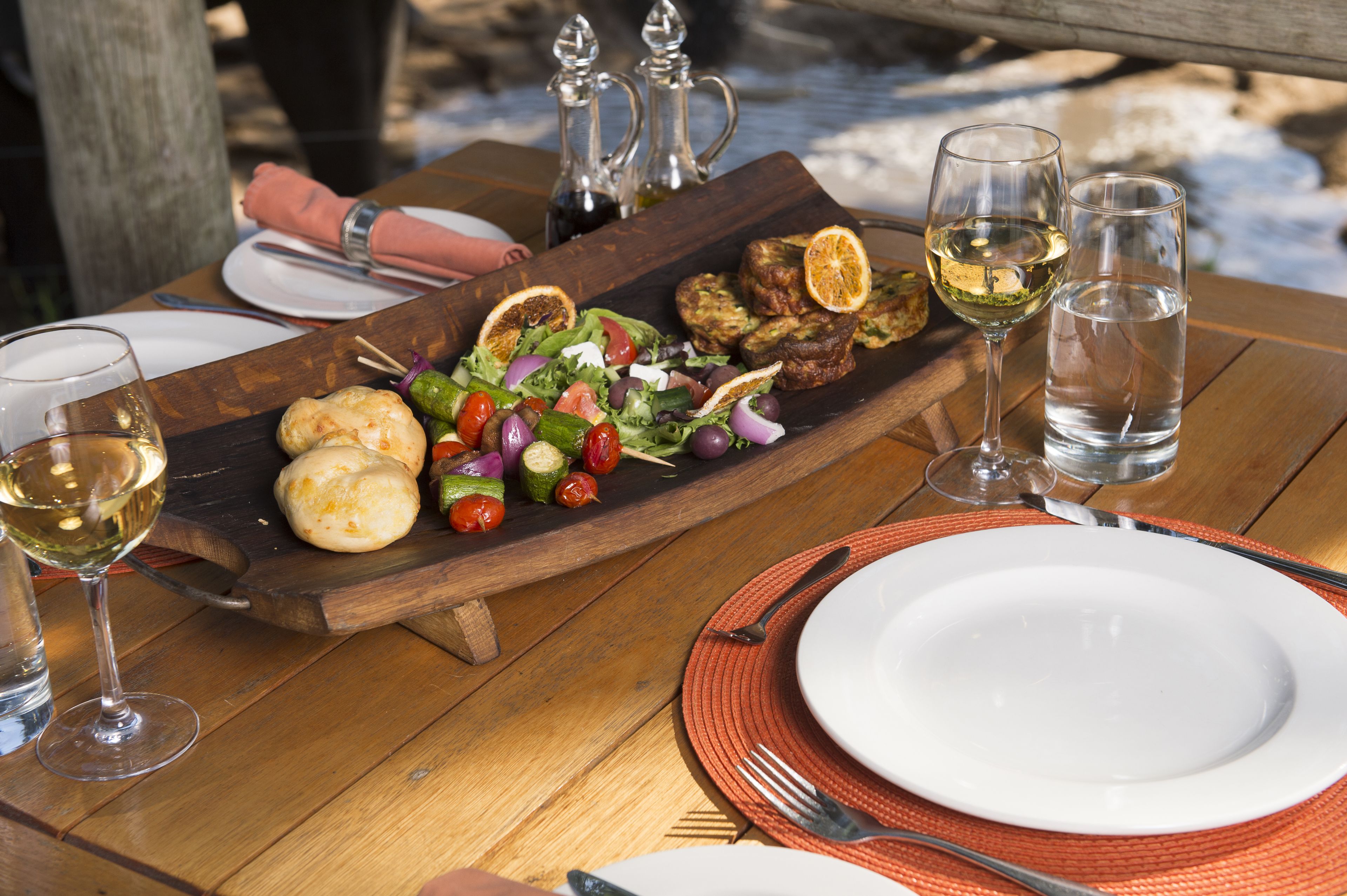 Une table en bois sur laquelle est posée une planche en bois servant de présentoir pour une salade, du pain et deux verres de vin blanc.