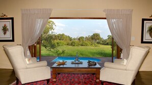 Una habitación con dos sillones blancos orientados hacia una mesa de centro de madera, una alfombra roja con diseño y vistas a una piscina y follaje exterior.