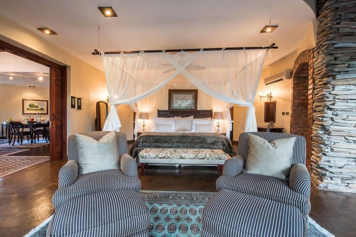 A bedroom with a canopy bed, two armchairs, a patterned rug, and a stone wall.