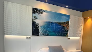Un dormitorio moderno con una cama blanca, una mesita de noche y una obra de arte en la pared que representa una escena costera.