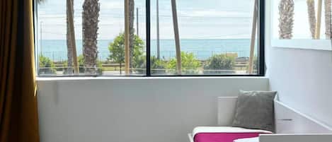 A bedroom with a bed, a window with a view of palm trees and the sea, and a small bedside table.