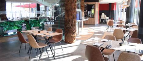 Un restaurant moderne doté d’un pilier central en bois, de rampes en verre et d’un escalier. Les tables et les chaises sont disposées pour accueillir les clients.