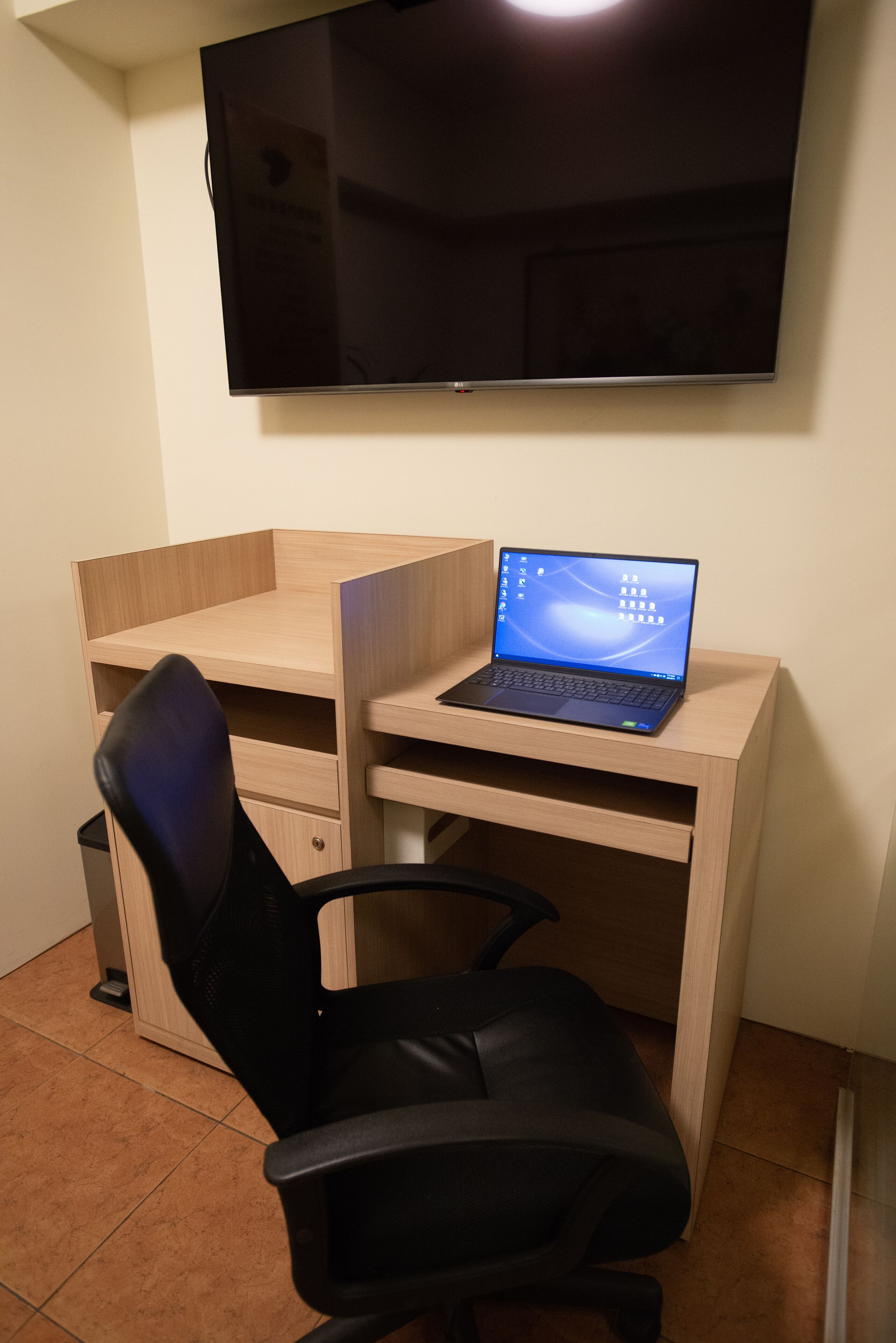 A small office space with a wooden desk, a black office chair, a laptop, and a flat-screen TV mounted on the wall.