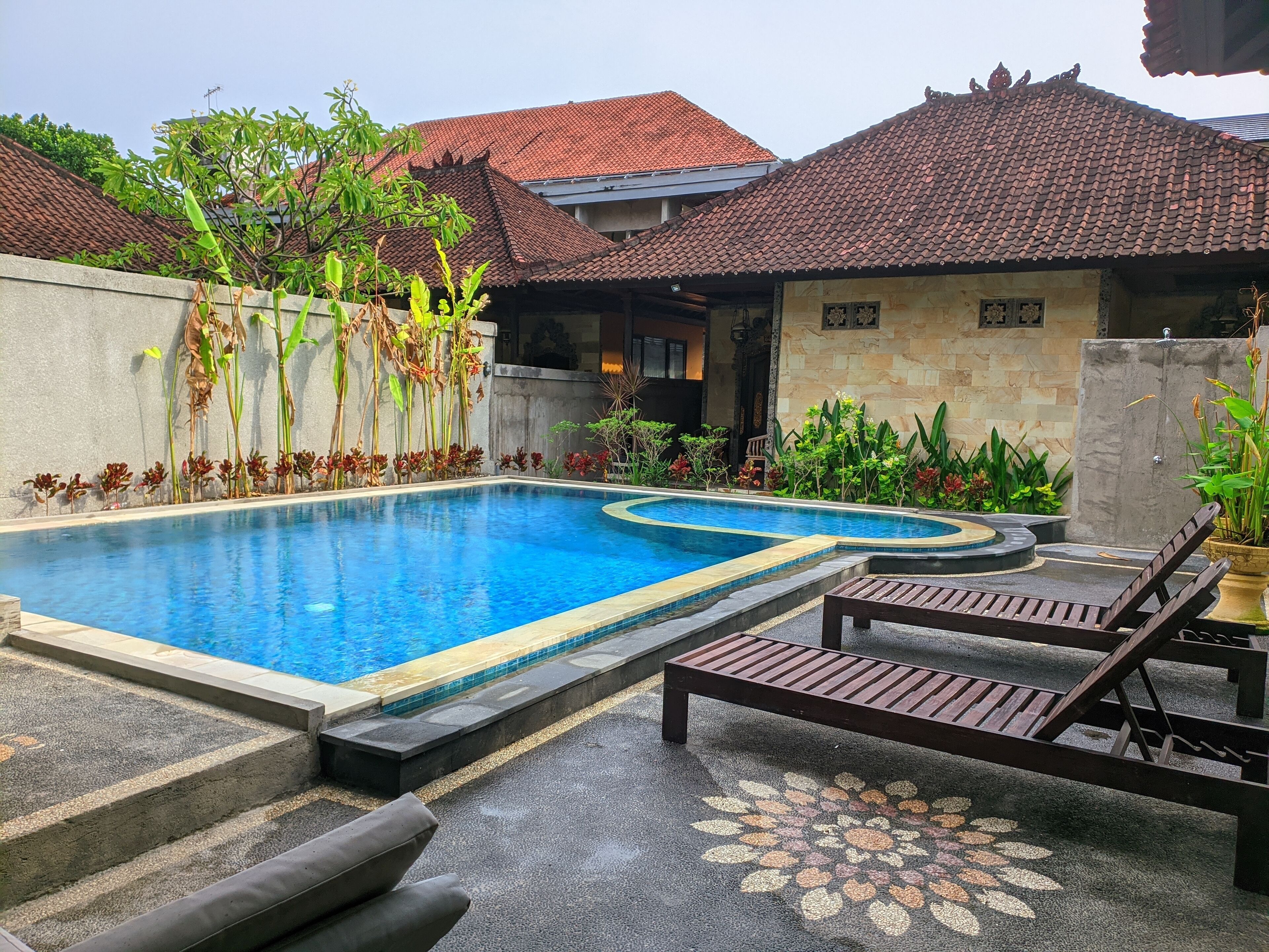 A swimming pool with a curved edge, surrounded by a concrete wall and various plants. There are lounge chairs and a decorative tile pattern on the ground.