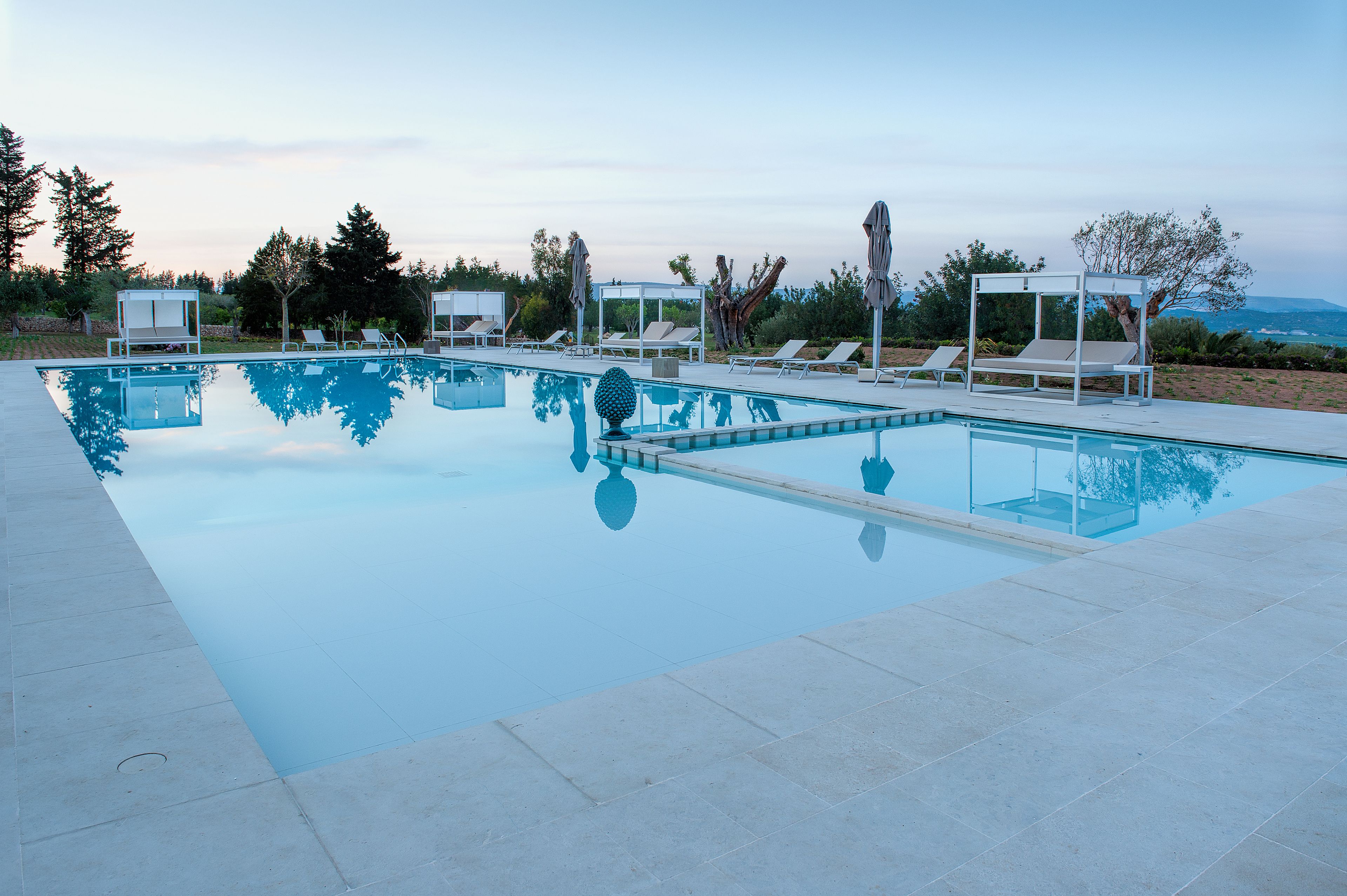A rectangular swimming pool with clear blue water, surrounded by white lounge chairs and umbrellas, set against a backdrop of trees and a clear sky.
