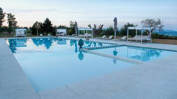 A rectangular swimming pool with clear blue water, surrounded by white lounge chairs and umbrellas, set against a backdrop of trees and a clear sky.
