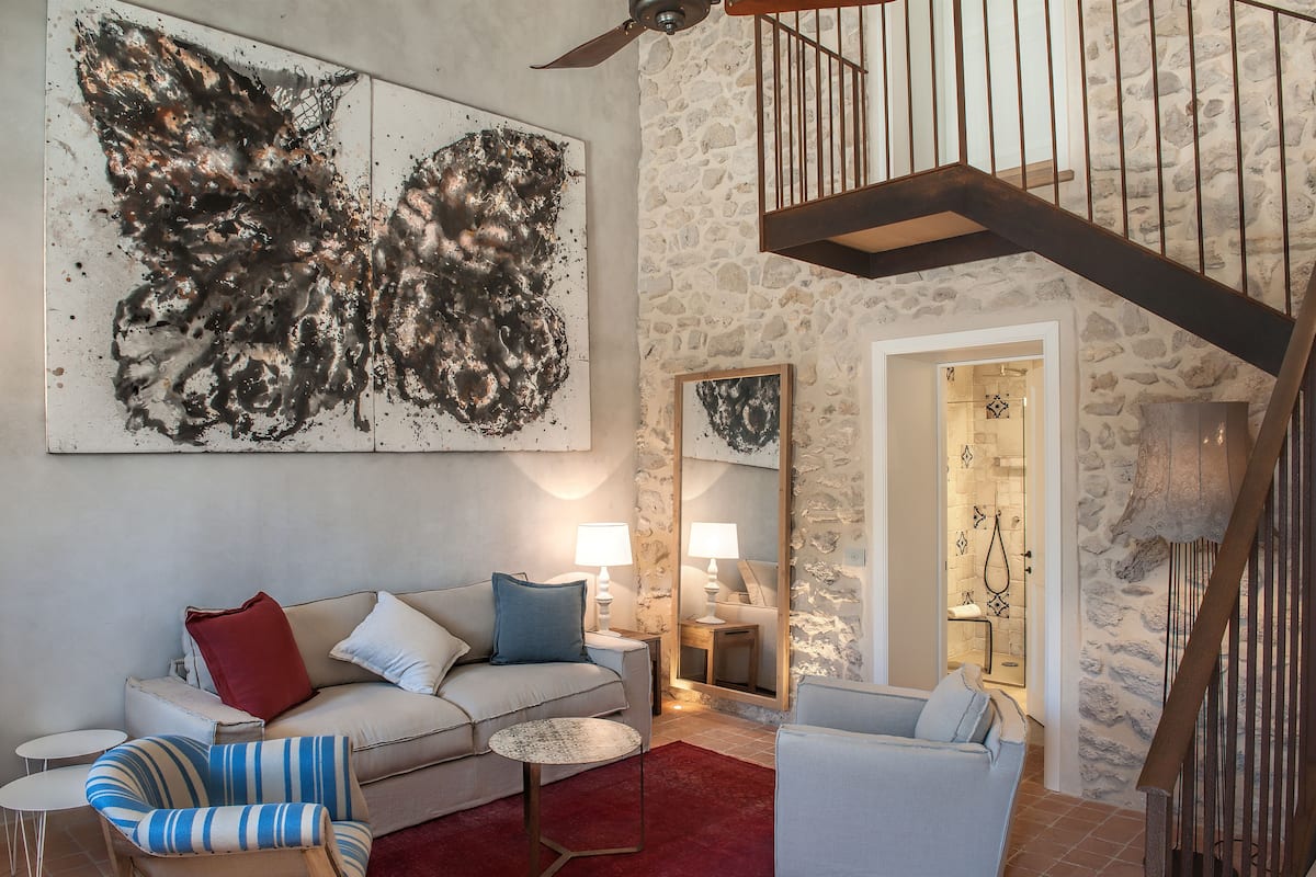 A living room with a stone wall, a staircase, a sofa, a chair, and abstract art on the wall.