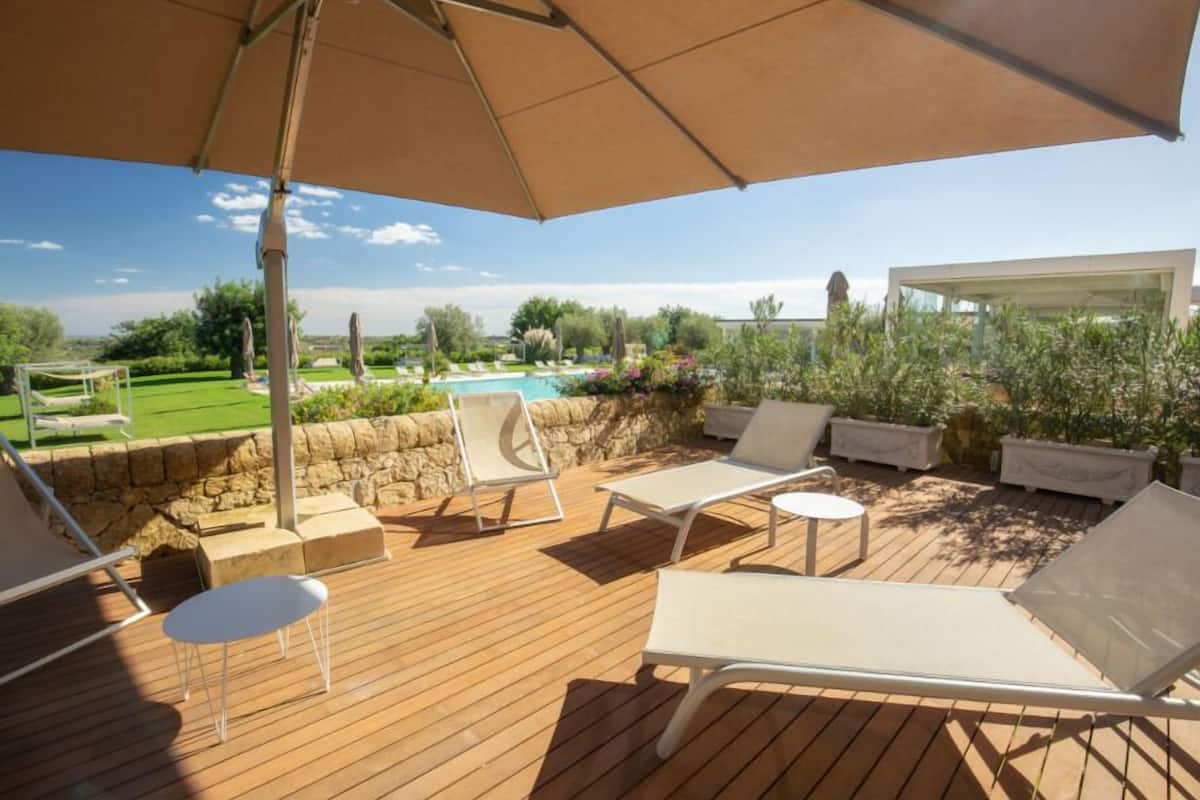 A wooden deck with lounge chairs and a table, a swimming pool, and a stone wall.
