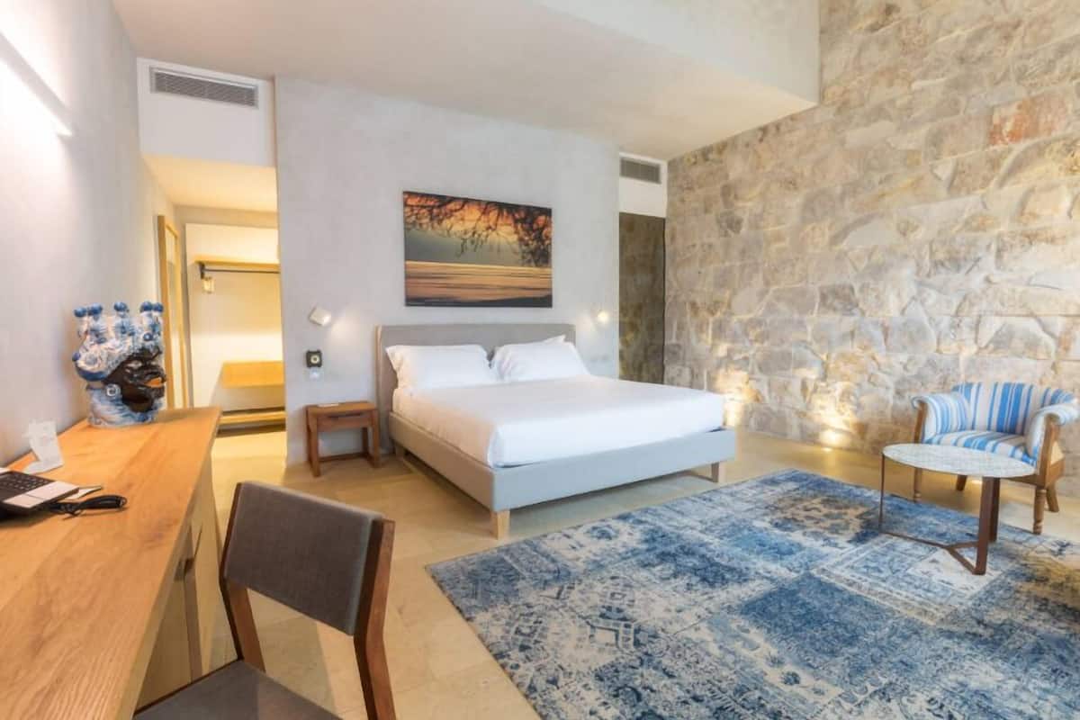 A bedroom with a bed, a desk, a chair, a rug, and a stone wall.