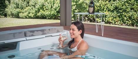 A person in a hot tub with a glass of white wine, surrounded by lush greenery and a table with a bottle.