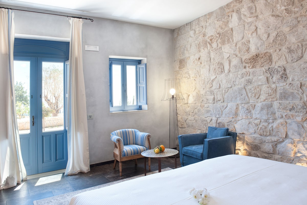 A bedroom with a stone wall, a bed, a chair, a table with fruit, and a lamp.