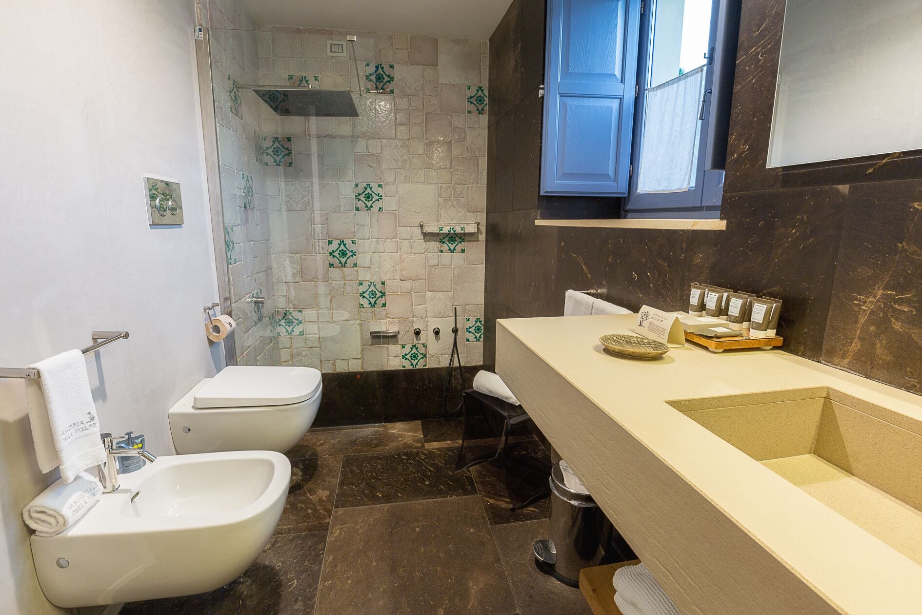 A bathroom with a white toilet, bidet, and sink. There is a shower area with patterned tiles, a towel rack, and a trash bin.