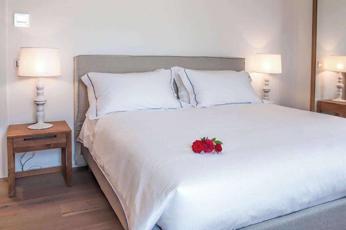 Una cama bien hecha con sábanas blancas y dos almohadas, una mesita de noche con lámpara y un espejo en la pared.