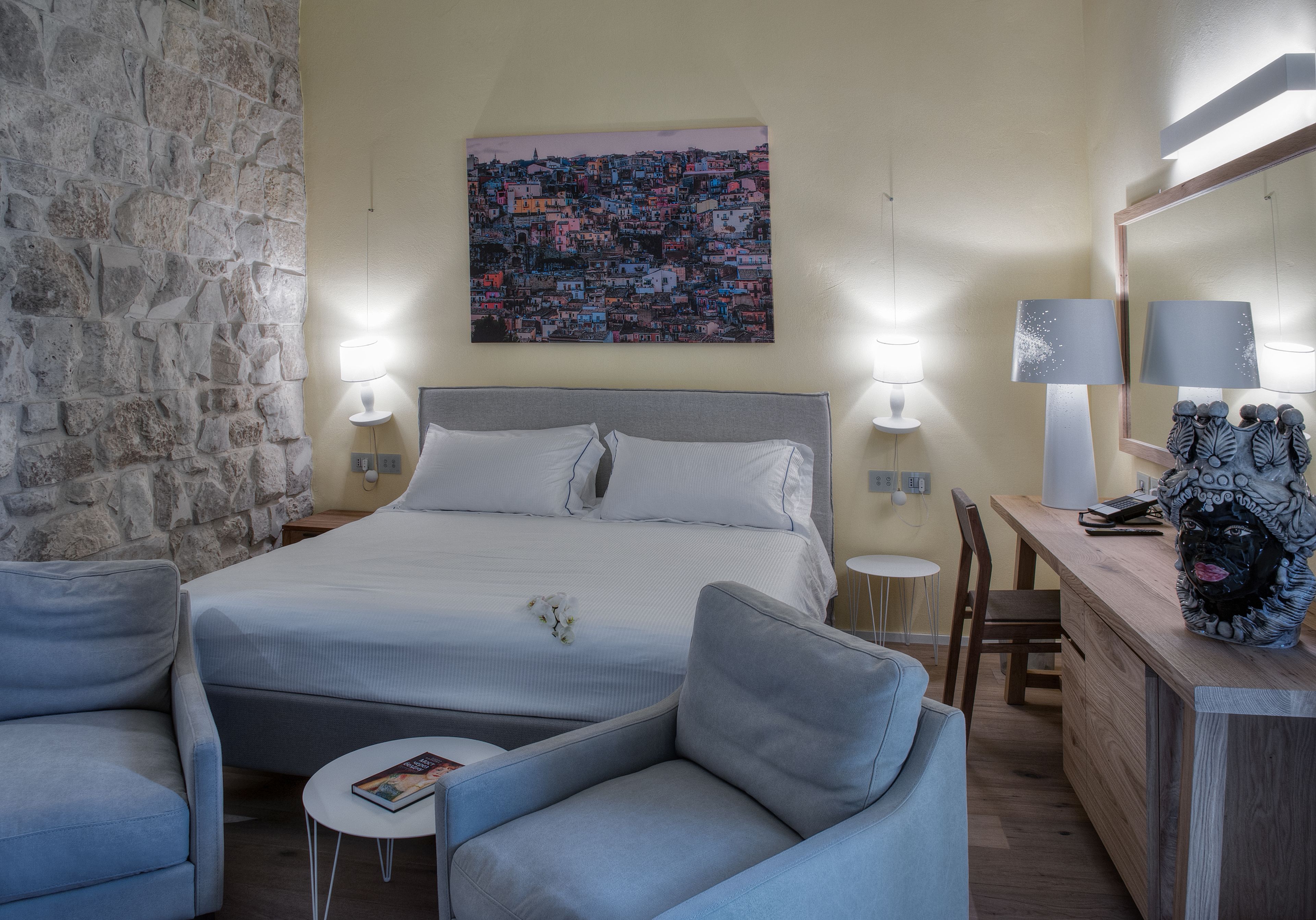 Un dormitorio con pared de piedra, una cama con ropa de cama blanca, dos lámparas de noche, una mesita redonda pequeña, una silla, un escritorio de madera y una escultura decorativa.