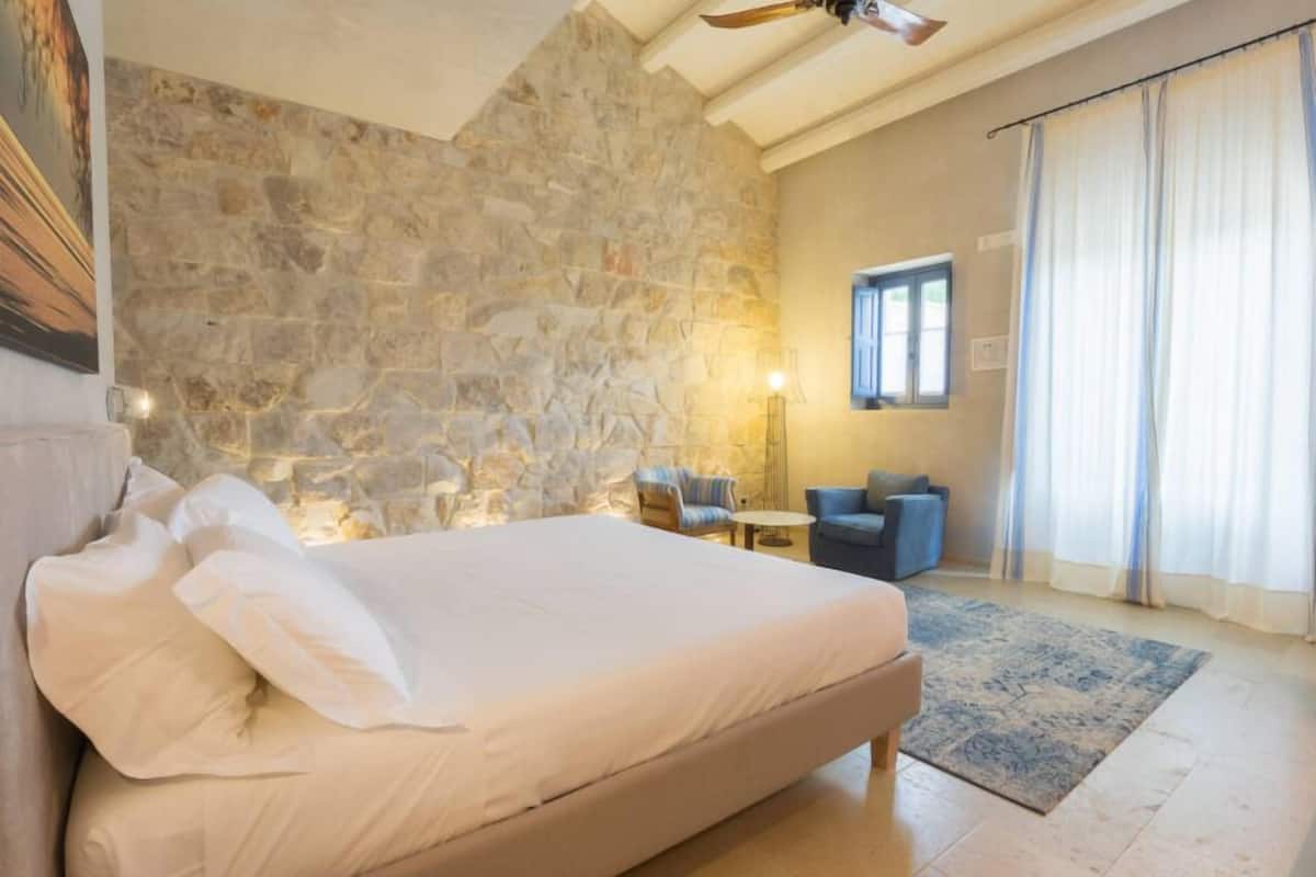 A bedroom with a stone wall, a bed, a chair, a table, and a window with curtains.