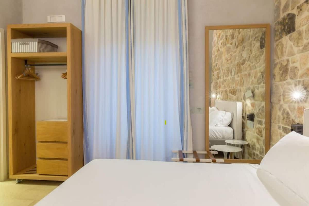 A bedroom with a wooden wardrobe, a bed, a stone wall, and a mirror.