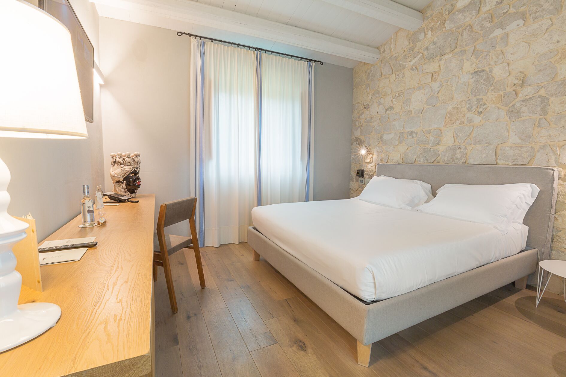 A hotel room with a large bed, a wooden desk, a chair, and a stone wall.