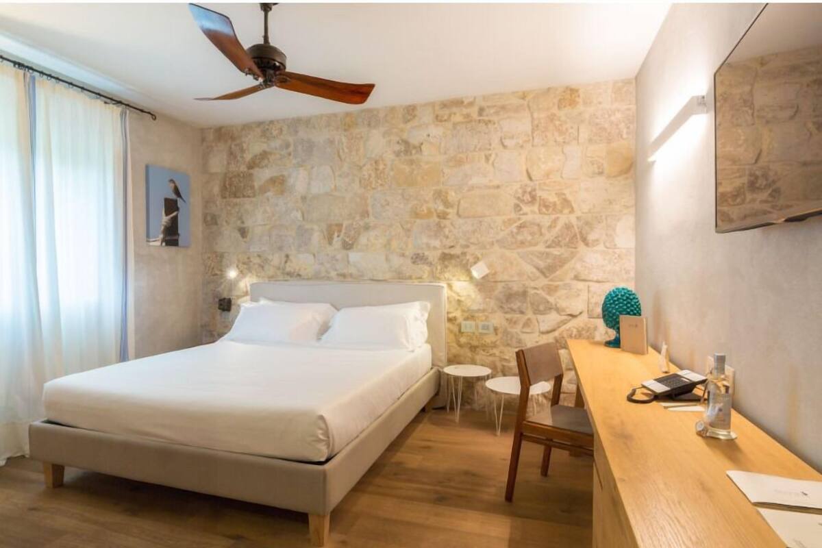 A bedroom with a bed, a desk, a chair, a ceiling fan, and a stone wall.