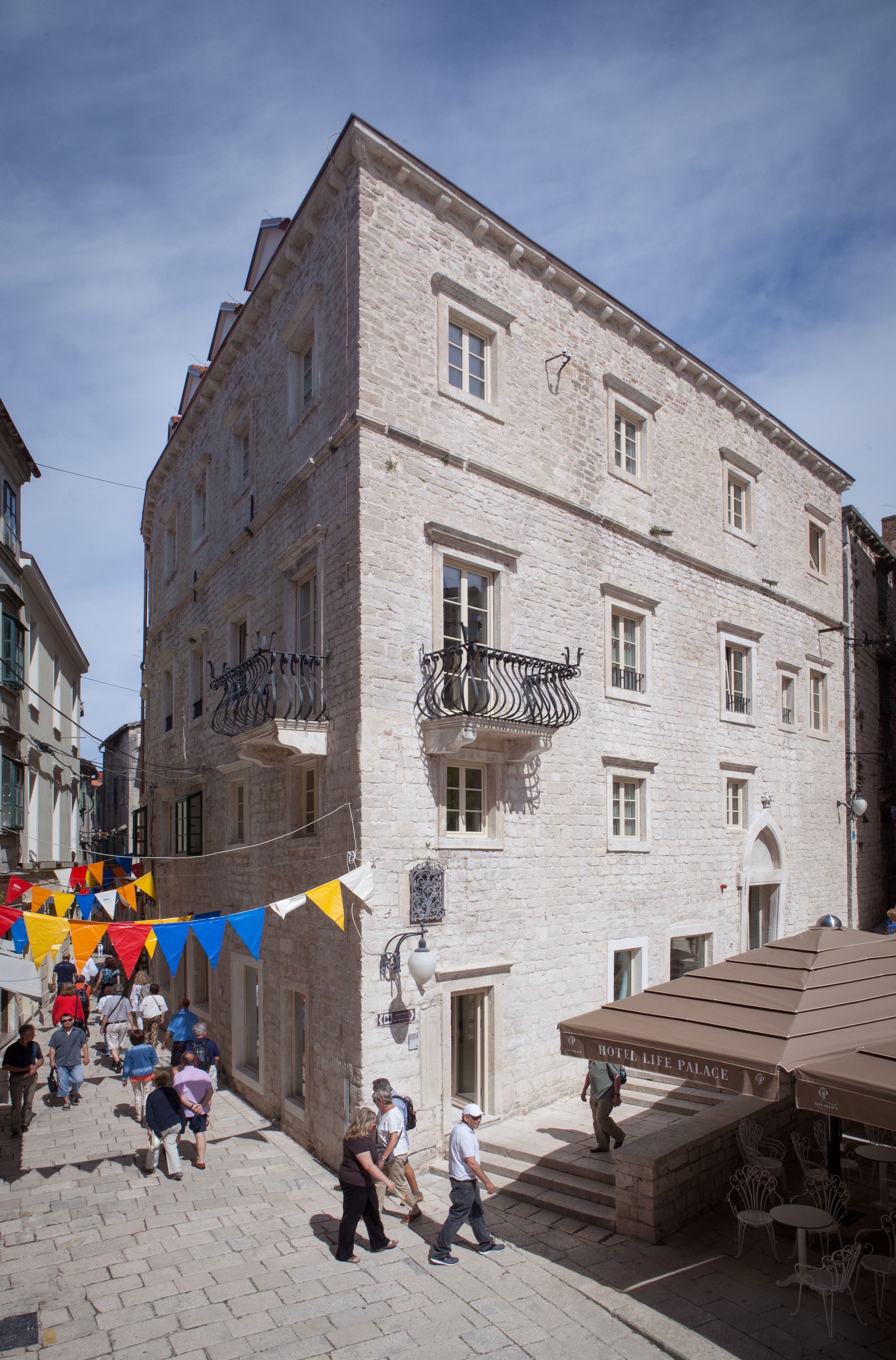 A stone building with multiple windows and balconies, a sign for Hotel Life Palace, and outdoor seating area.
