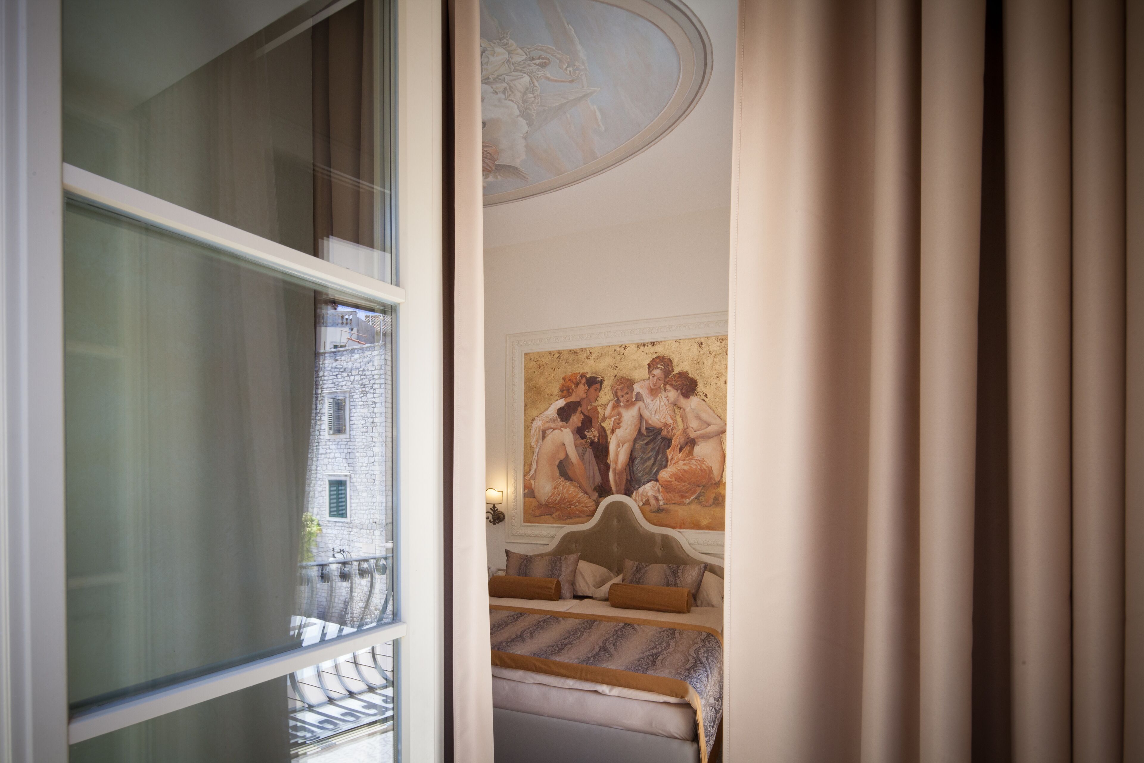 A room with a bed, a painting, and a view of buildings outside.