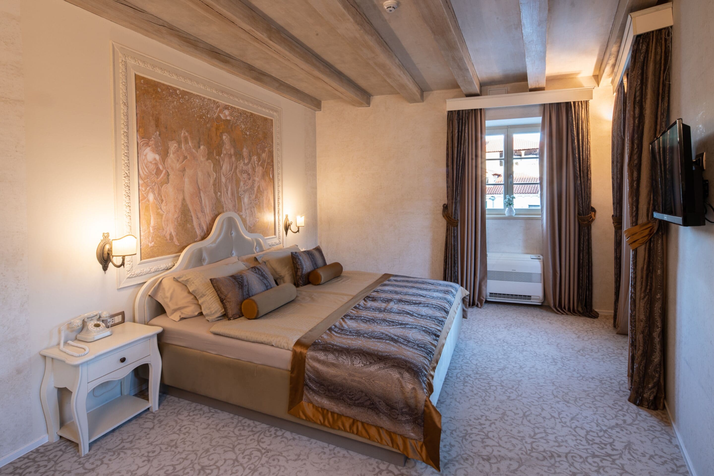 Ein Hotelzimmer mit einem großen Bett, einem Nachttisch mit Telefon, einem Bild an der Wand und Blick durch eine Tür nach draußen.