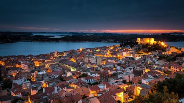 Une ville côtière au crépuscule, avec des bâtiments illuminés et un château imposant qui se dresse sur une colline.