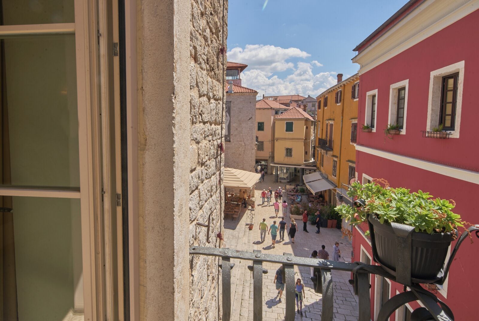 A narrow street with buildings on either side, a pedestrian area, and a balcony with a potted plant.