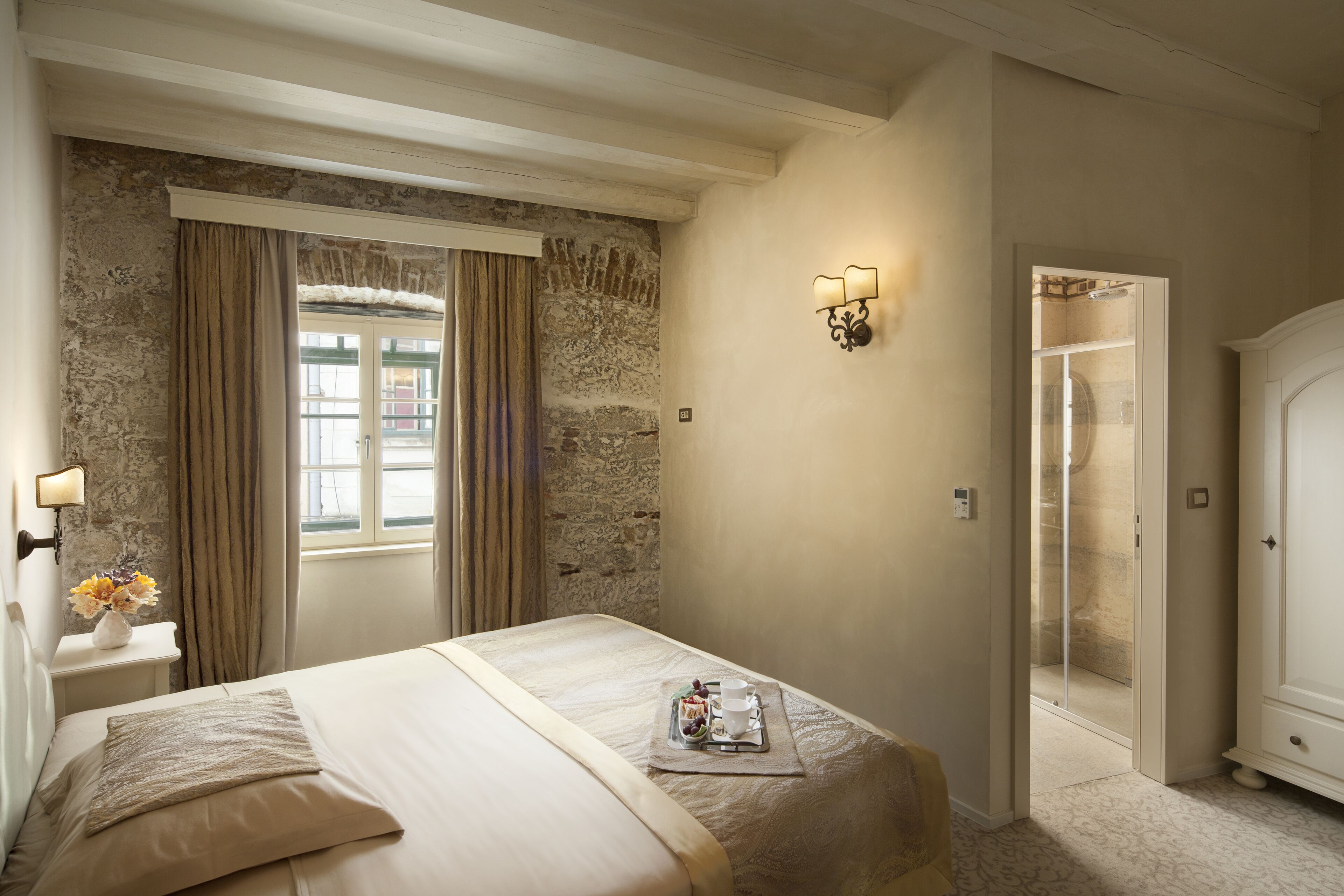 A bedroom with a stone wall, a bed with a tray of breakfast, a nightstand with a vase of flowers, and a bathroom with a shower.
