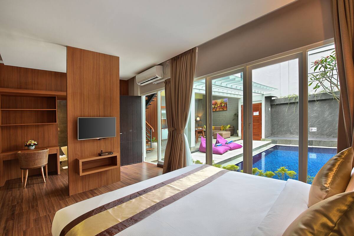 A modern hotel room with a large bed, a TV, a desk, and a view of a swimming pool.