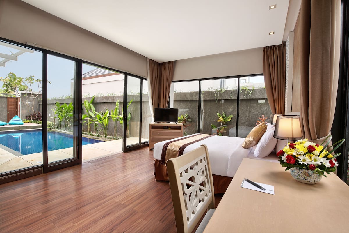 A hotel room with a large bed, a desk with a lamp, a flower vase, and a view of a swimming pool.