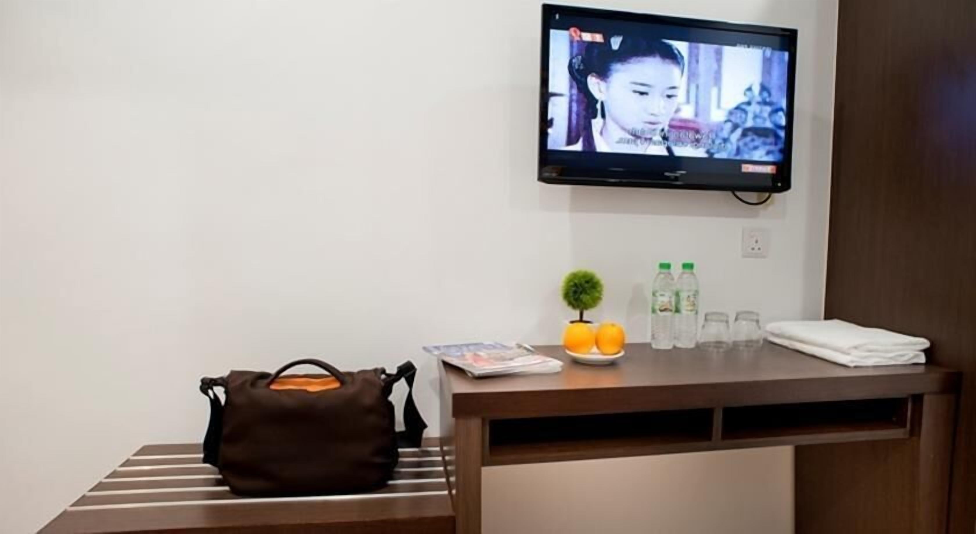 Habitación de hotel con televisor montado en la pared, un escritorio de madera con un bolso, una pequeña planta en maceta, un plato con naranjas y dos botellas de agua.