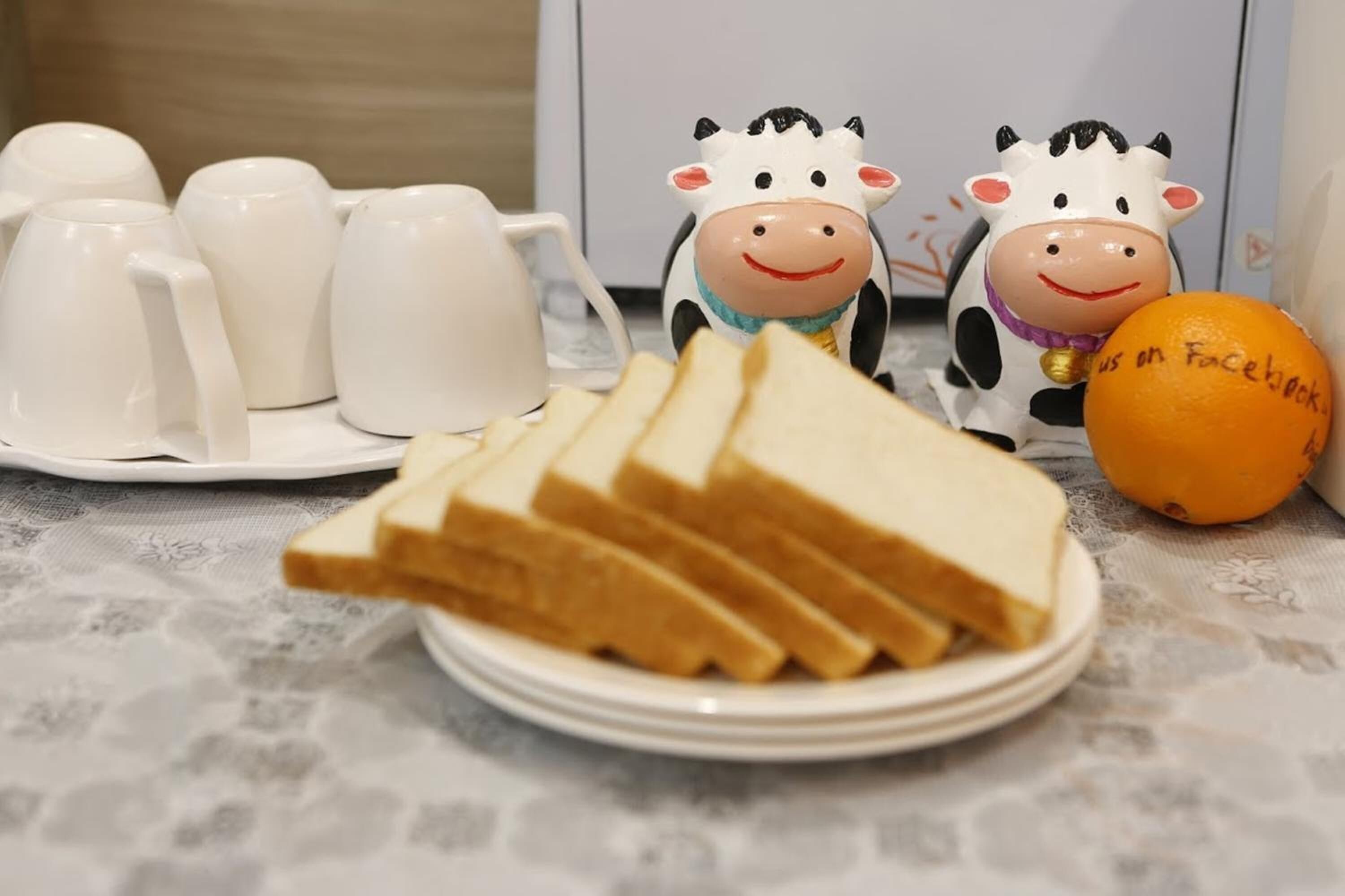Een bord met sneetjes toast, een hele sinaasappel en twee koeienbeeldjes op een tafel met witte kopjes en schotels.
