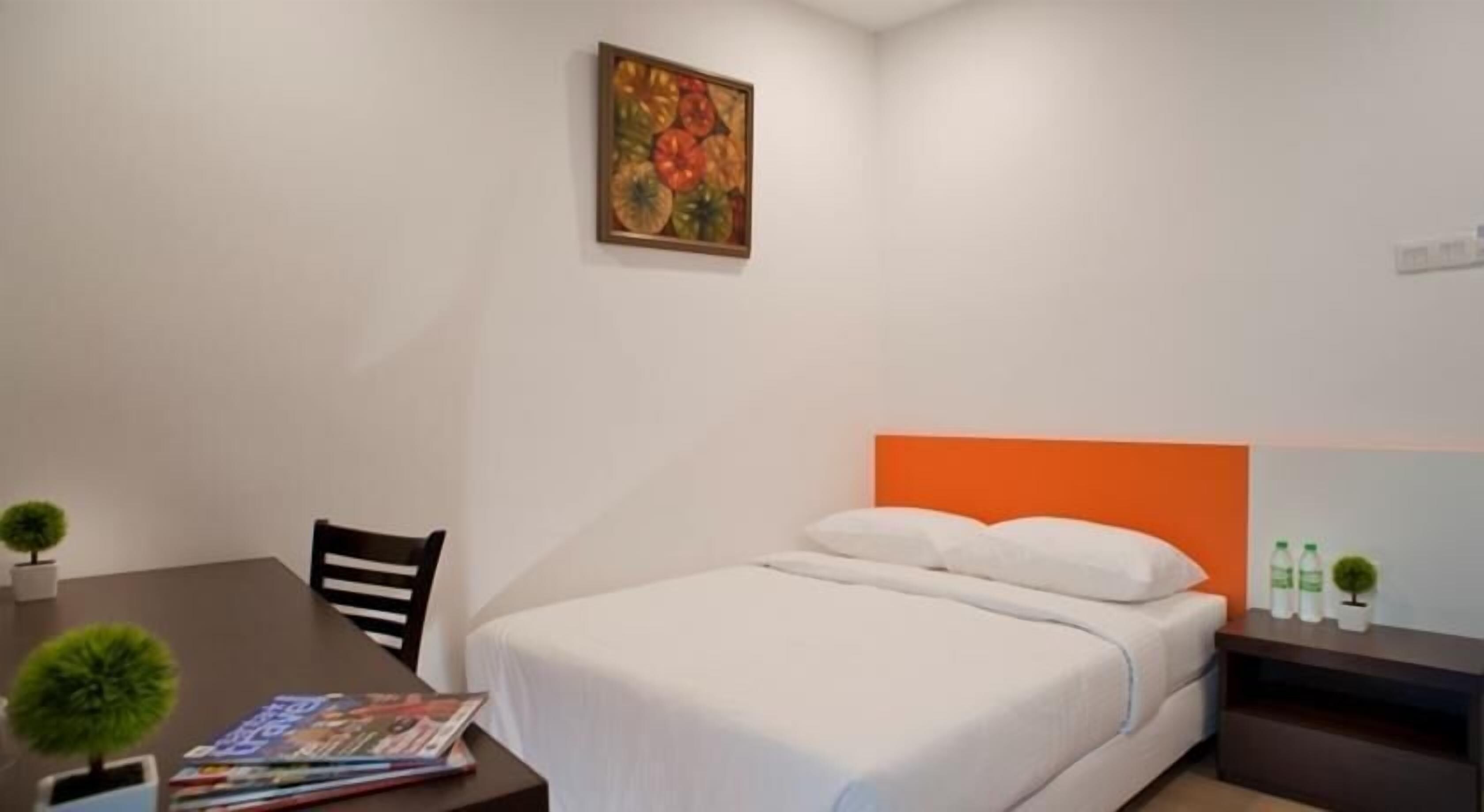 A hotel room with a bed, a table with magazines, a chair, a small plant, and a painting on the wall.
