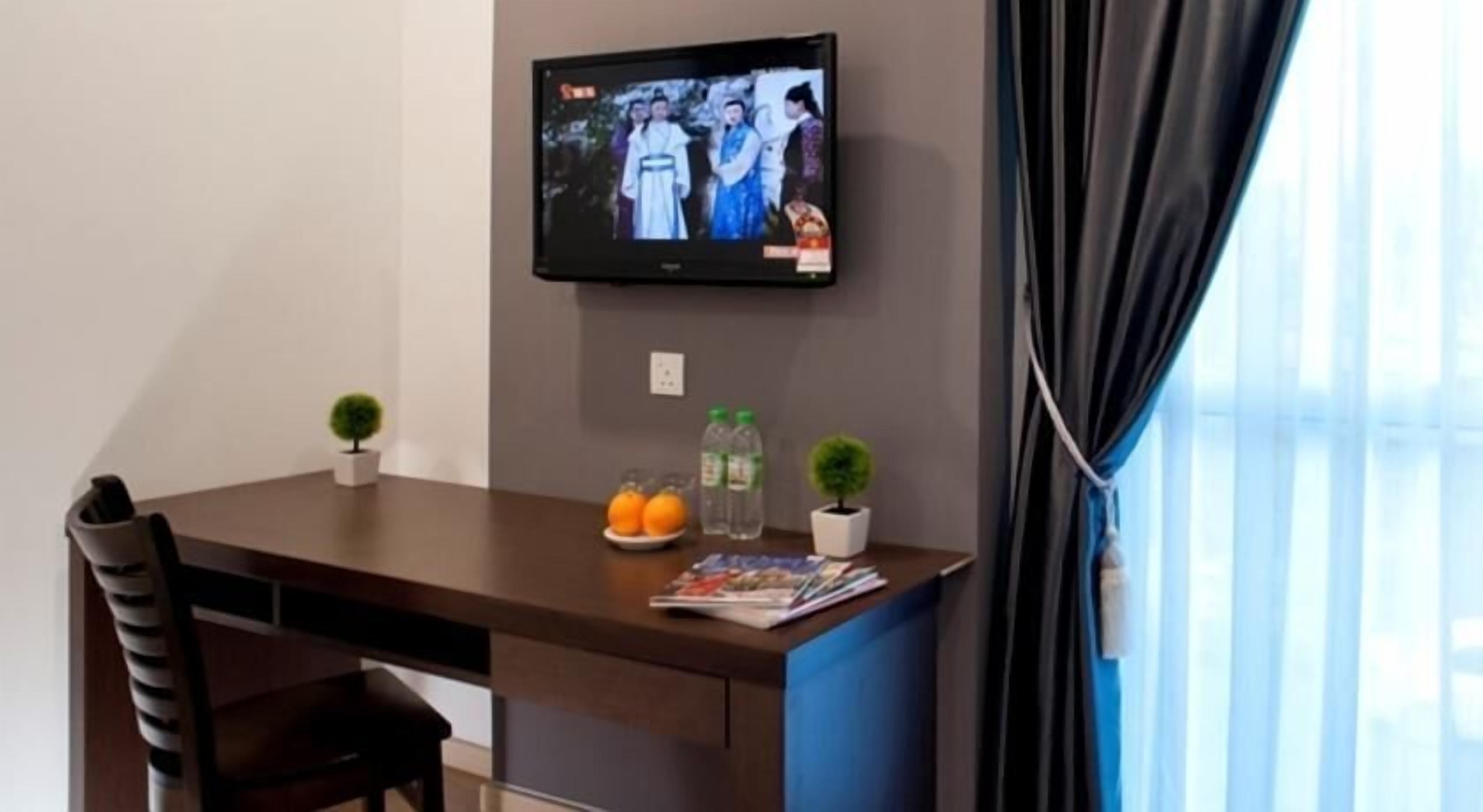 A hotel room with a TV mounted on the wall, a small table with a chair, two potted plants, a plate with oranges, and a magazine.