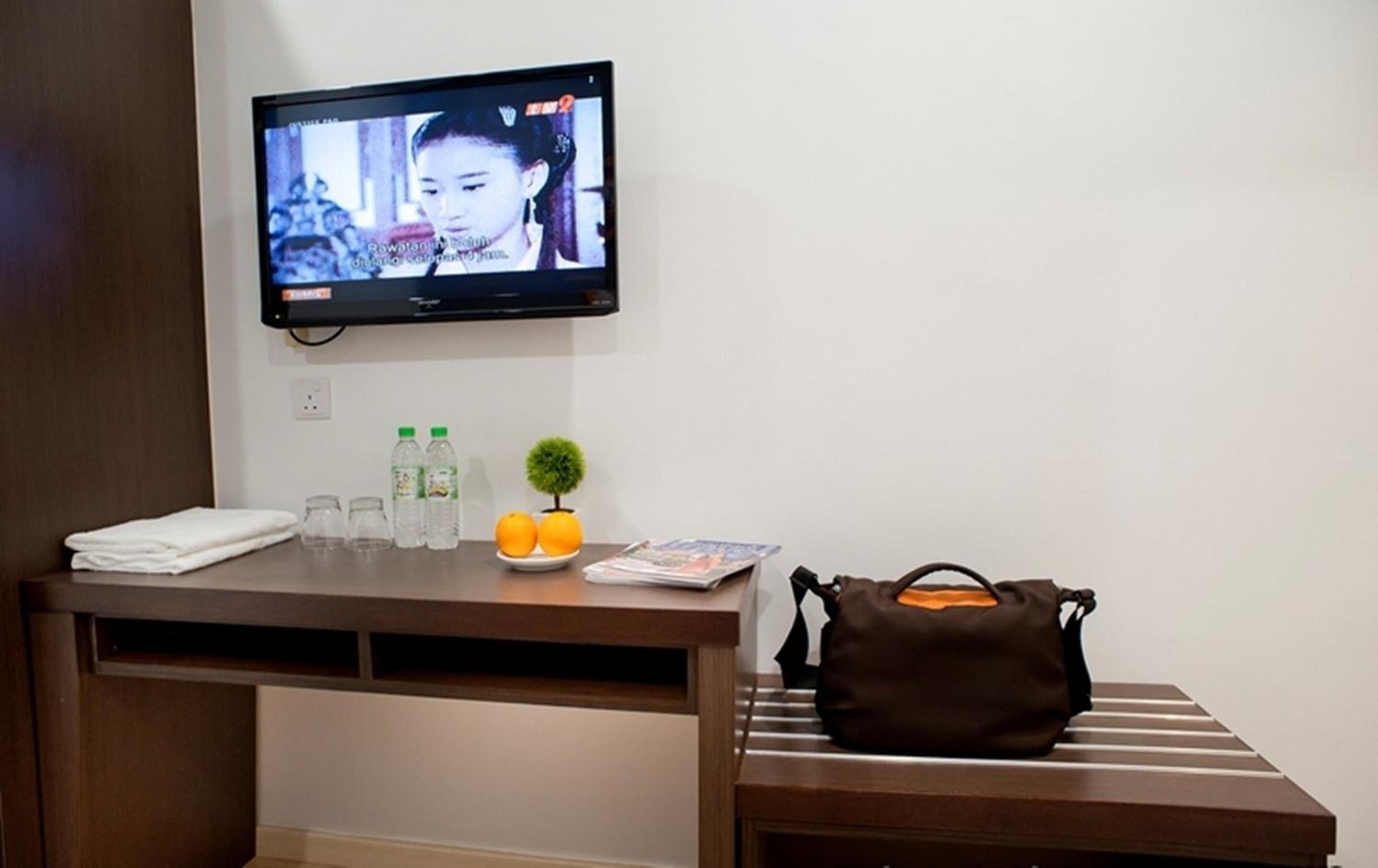 Een hotelkamer met een aan de muur bevestigde televisie, een bureau met een laptop, een handtas, een kleine plant en een bord met mandarijnen.