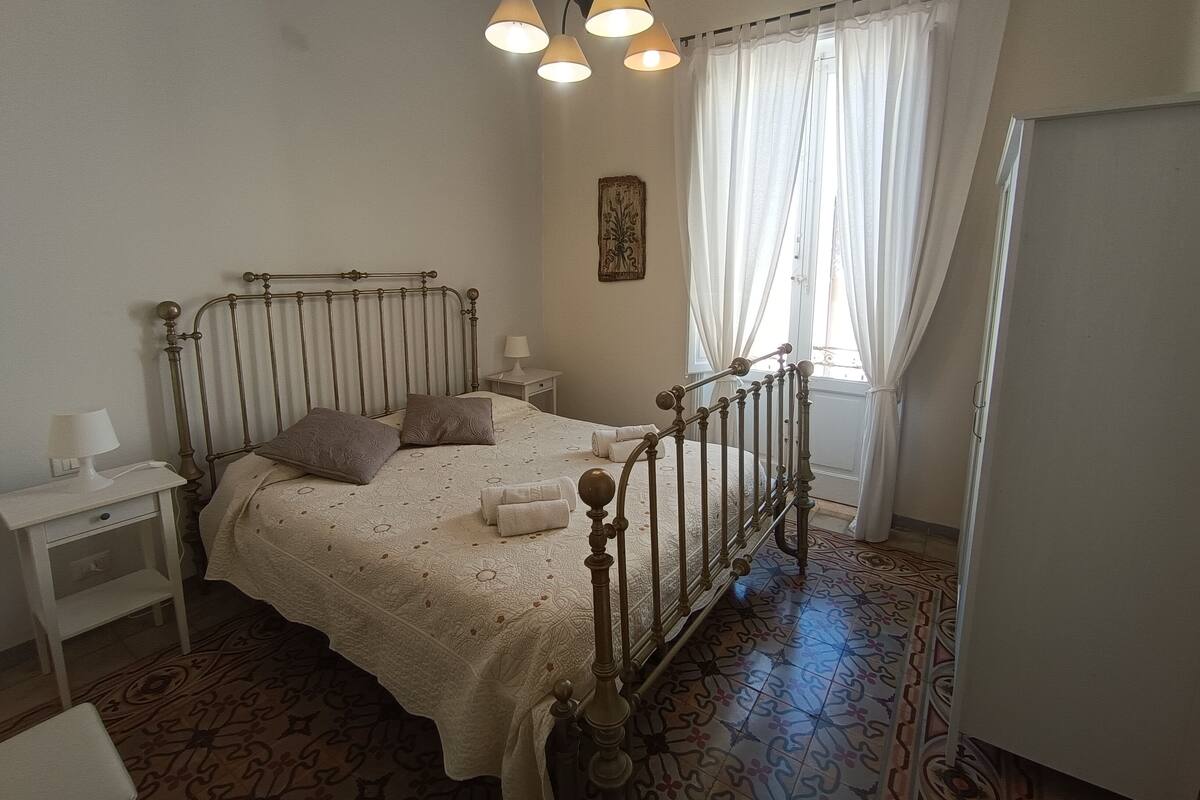 A bedroom with a bed, bedside tables, a lamp, a window with curtains, and a patterned floor.