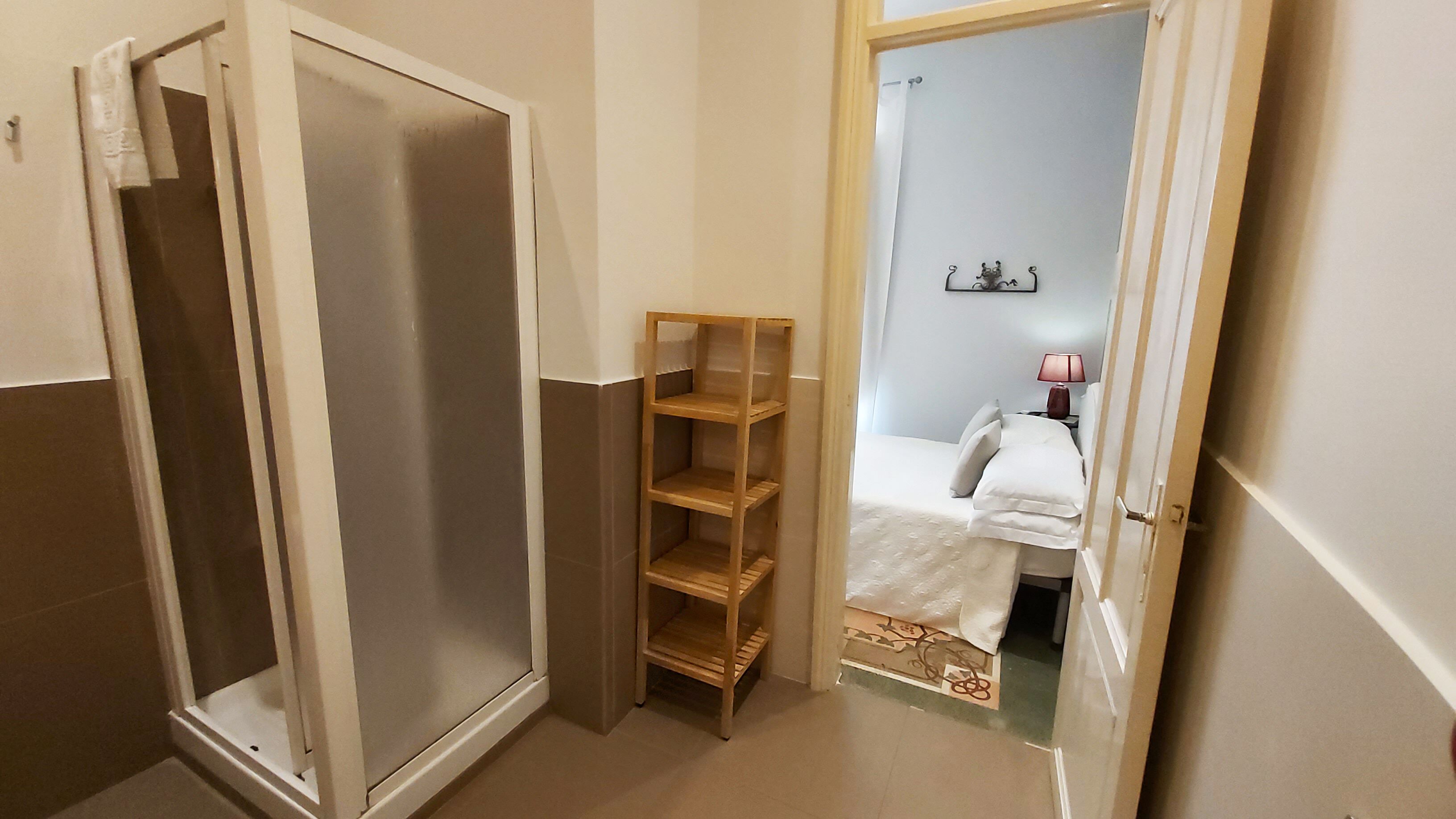 A narrow hallway with a wooden shelf unit, a door leading to a bedroom, and a small bedside table with a lamp.