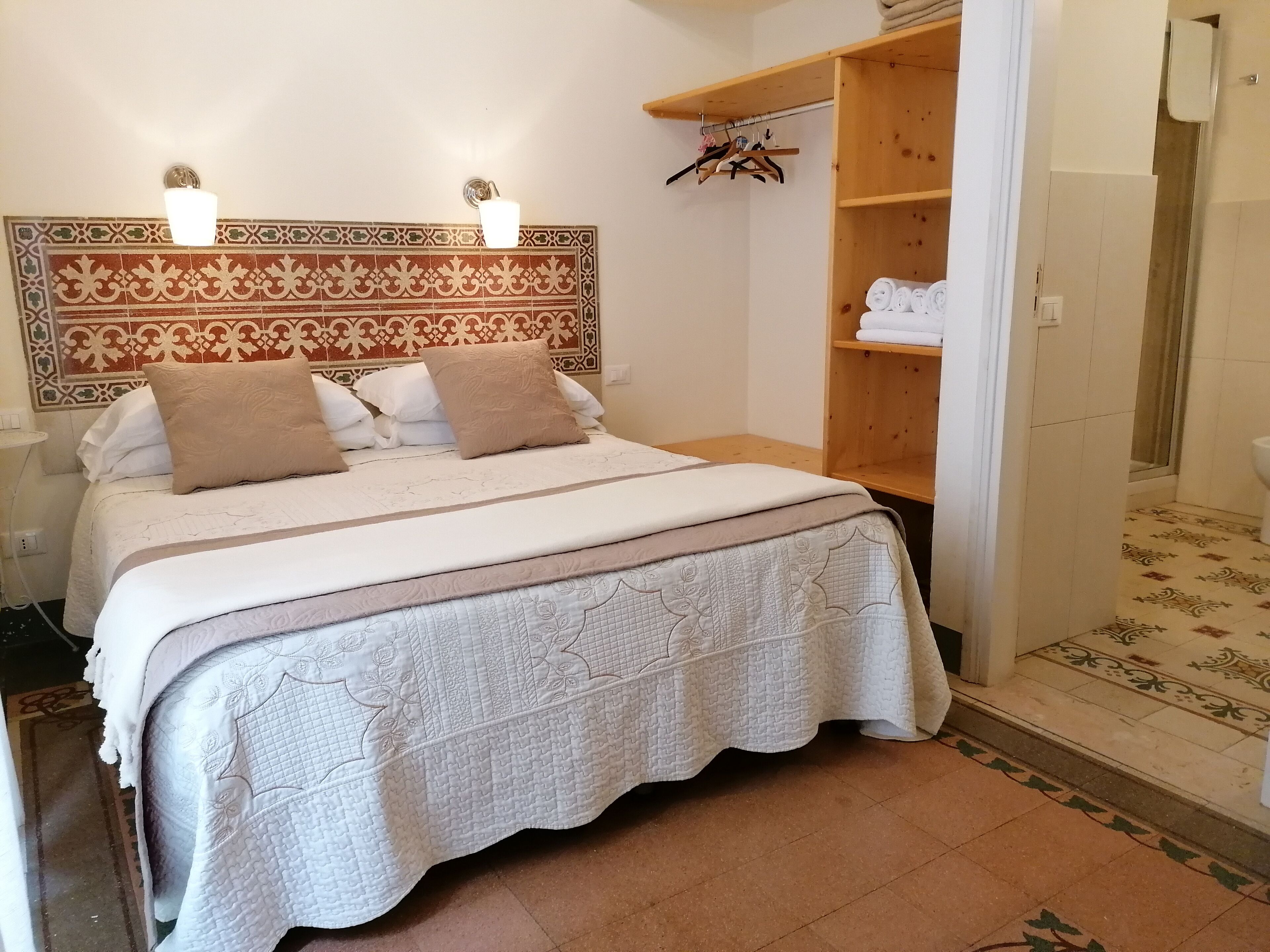 A neatly made bed with a patterned headboard, a wooden shelf with folded towels, and a tiled floor.