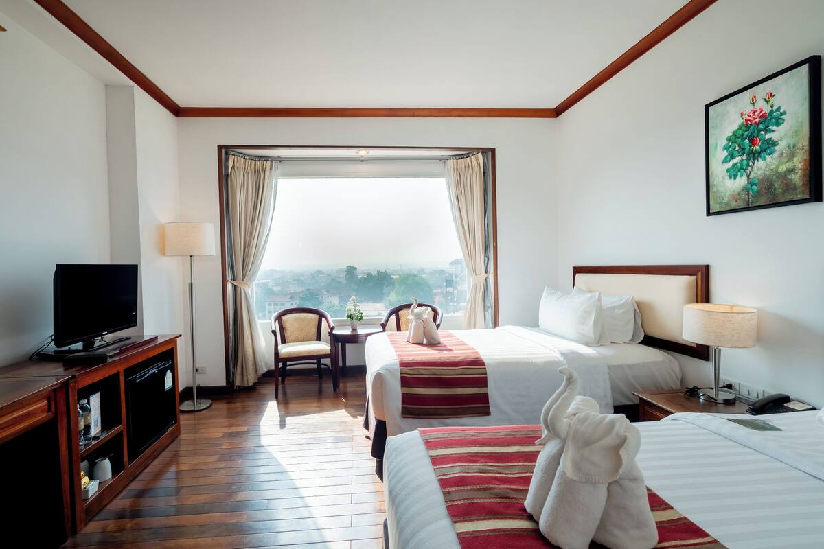 A hotel room with two beds, a TV, a desk, a chair, and a view of the city.