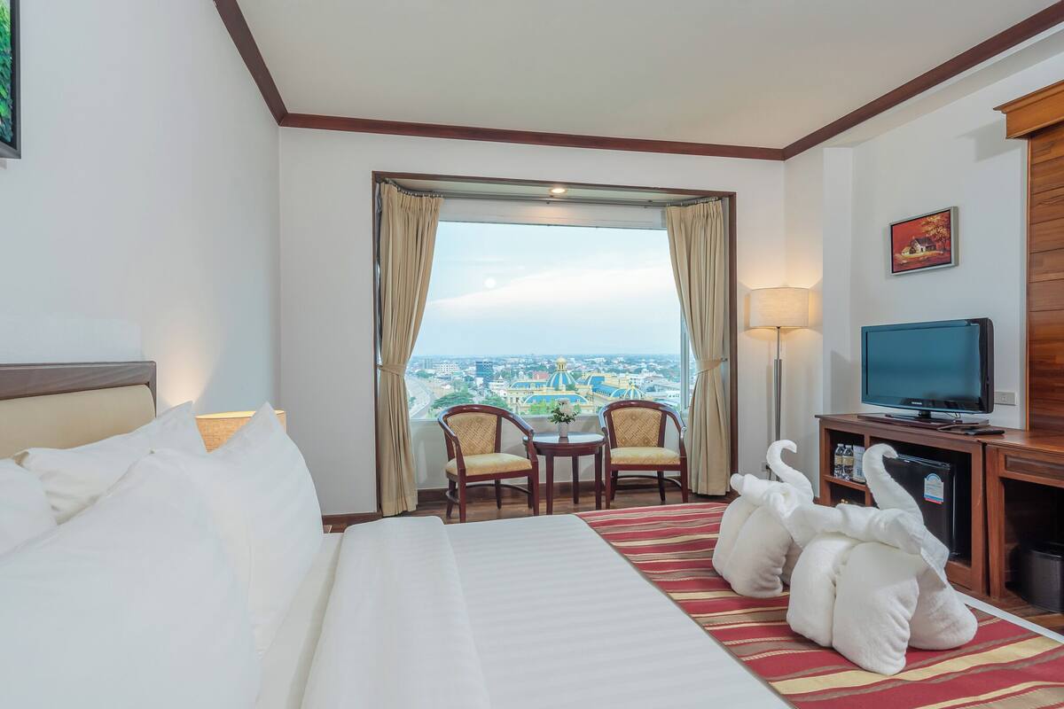 A hotel room with a bed, two chairs, a TV, and a large window with a view of a cityscape.