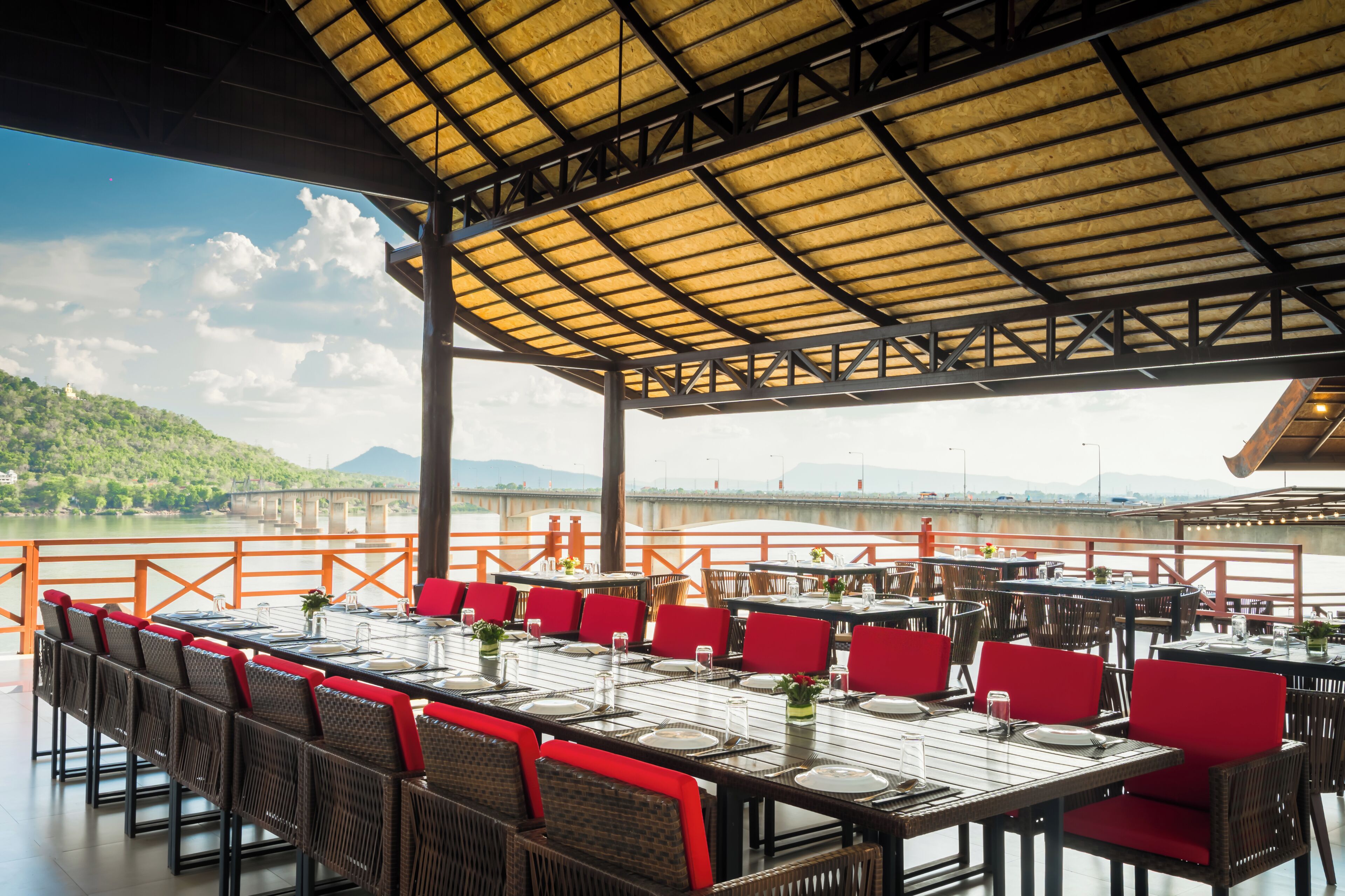 An outdoor dining area with tables and chairs, a covered roof, and a view of a bridge and mountains.