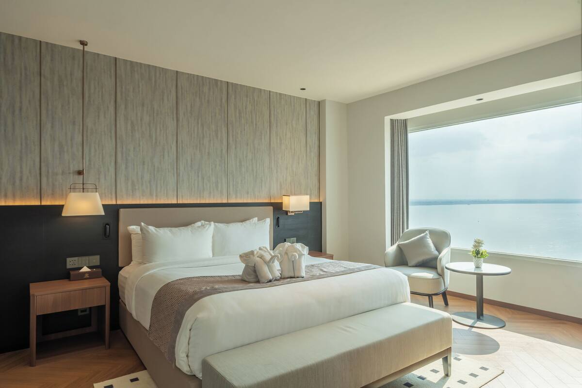 A modern hotel room with a large bed, bedside tables, a seating area, and a view of the sea.