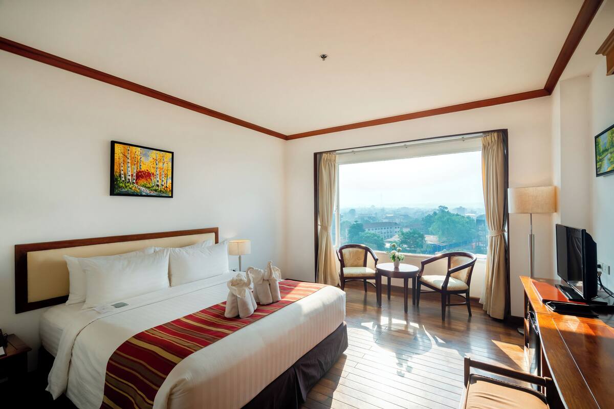 A hotel room with a bed, a painting, a television, a desk, and a view of the city.