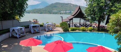 A swimming pool with lounge chairs and umbrellas, surrounded by greenery and a view of a lake and mountains.