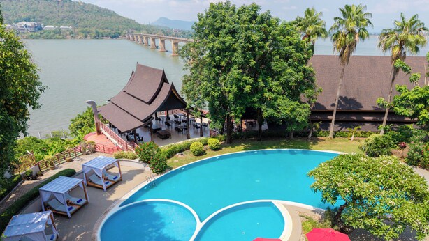 Une piscine avec des chaises longues et des parasols, un restaurant doté d’une terrasse, et un pont en arrière-plan.