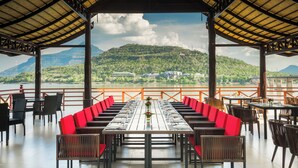 Un espace extérieur aménagé pour les repas, avec des tables et des chaises, une vue sur une montagne et un plan d’eau, et une structure couverte avec un toit.