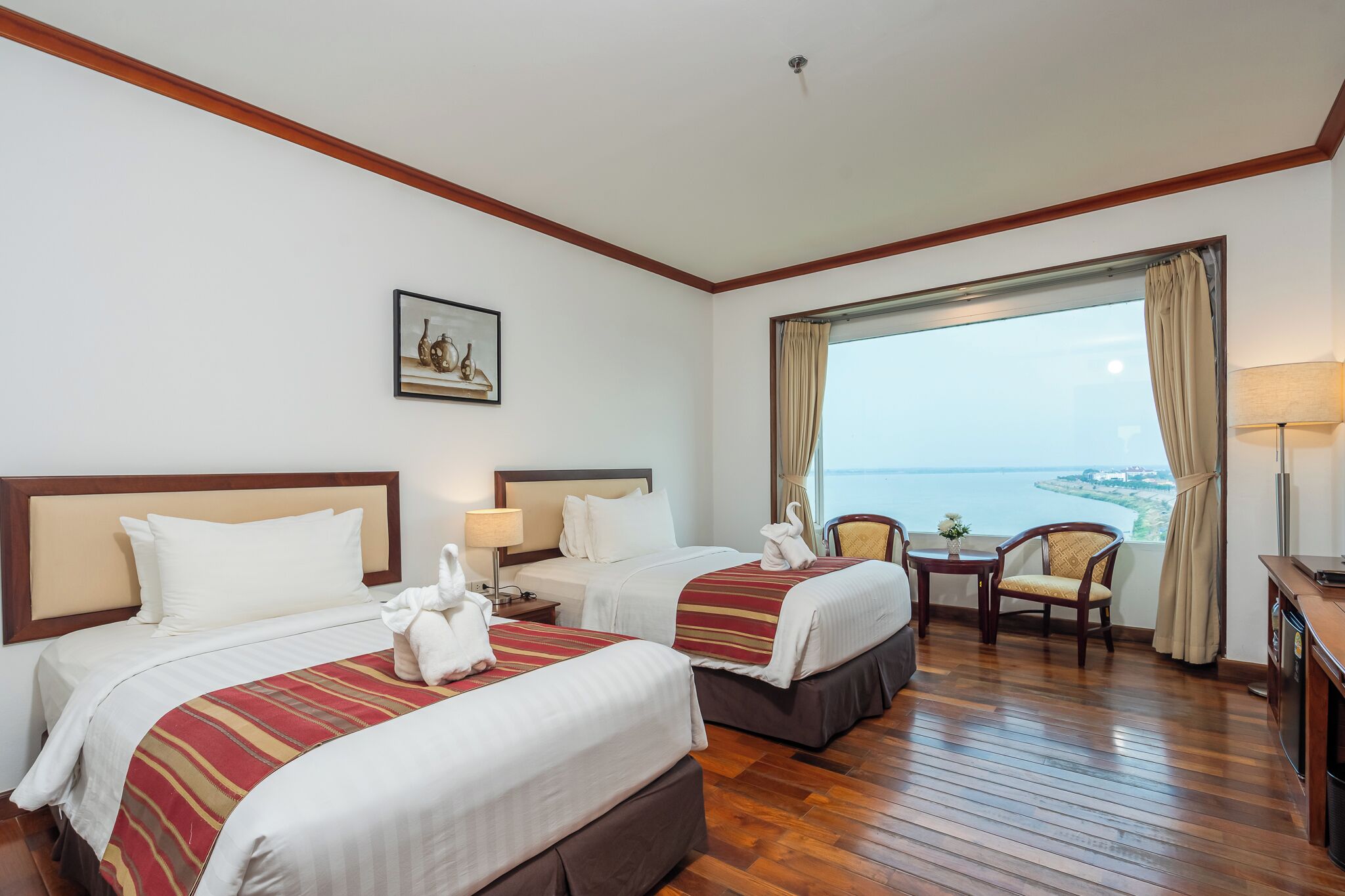 A hotel room with two beds, a wooden floor, a view of the sea, a painting on the wall, and a small table with a lamp.