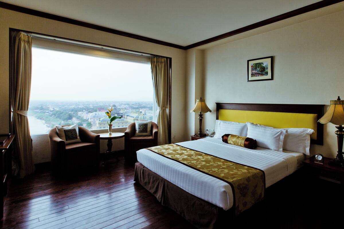 A hotel room with a large bed, two bedside tables with lamps, a small table with a vase, a chair, and a view of a cityscape through a large window.