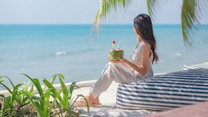 A person sitting on a striped cushion by the ocean, holding a coconut drink.