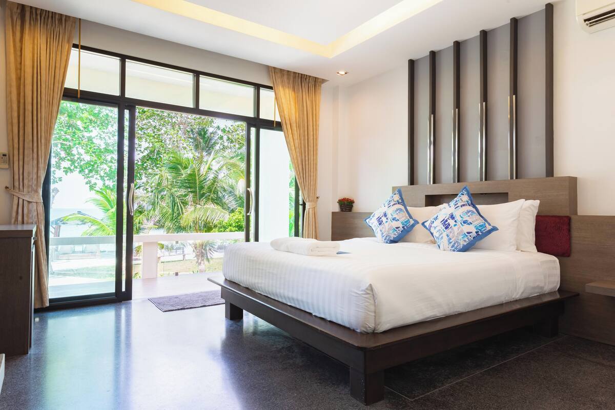 A modern hotel room with a large bed, a nightstand, a window with curtains, and a view of greenery outside.